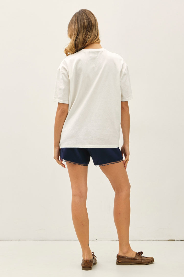 A woman shown from the back wearing a white short-sleeve t-shirt, navy blue shorts, and brown boat shoes against a plain white studio background.