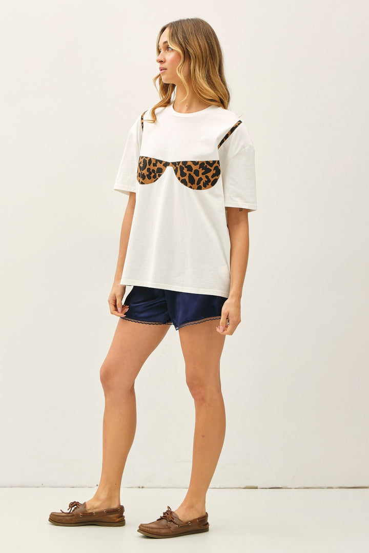 A woman modeling a white short-sleeve t-shirt with a leopard-print bra graphic on the front, paired with navy blue denim shorts and brown boat shoes, against a plain white studio background.