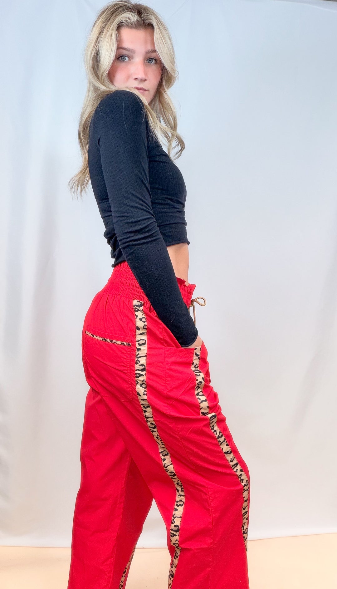 Woman shown from side view wearing black long sleeve top with red windbreaker pants with cheetah print stripes on plain background