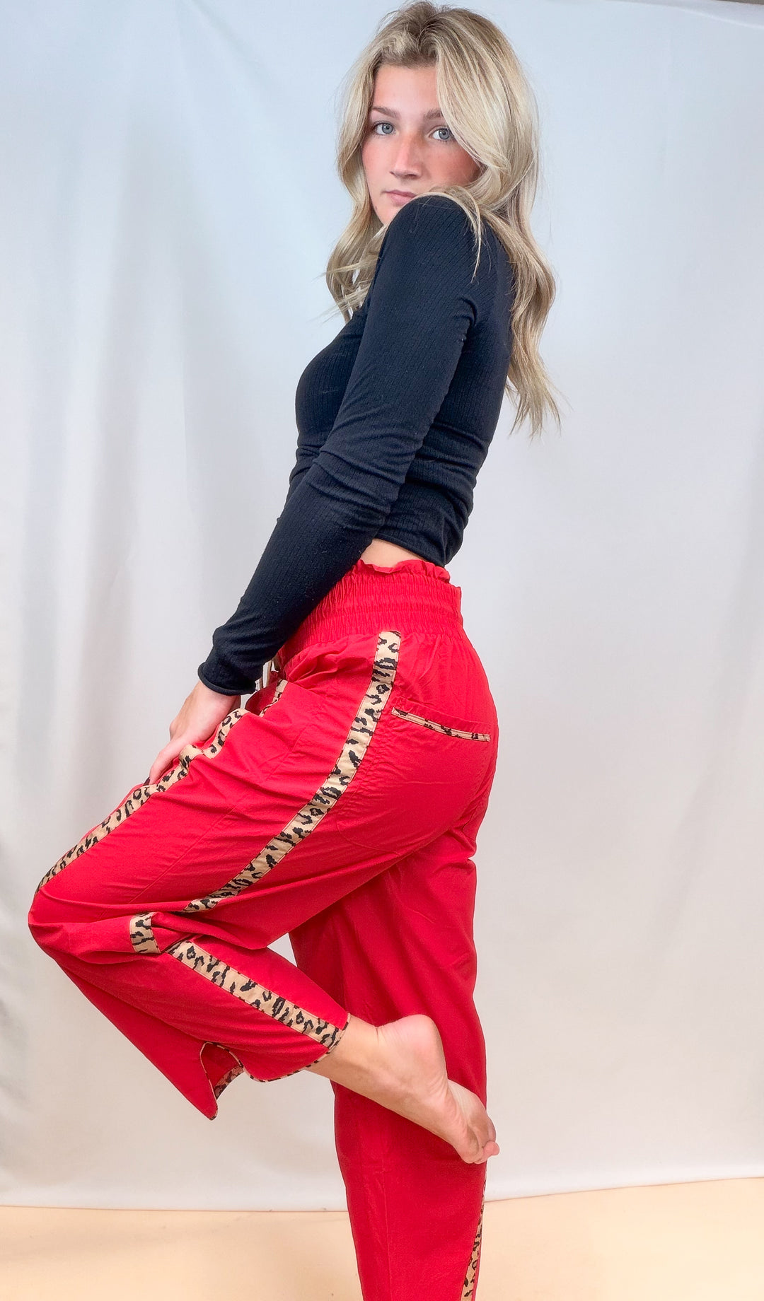 Woman shown from side view wearing black long sleeve top with red windbreaker pants with cheetah stripes on plain background