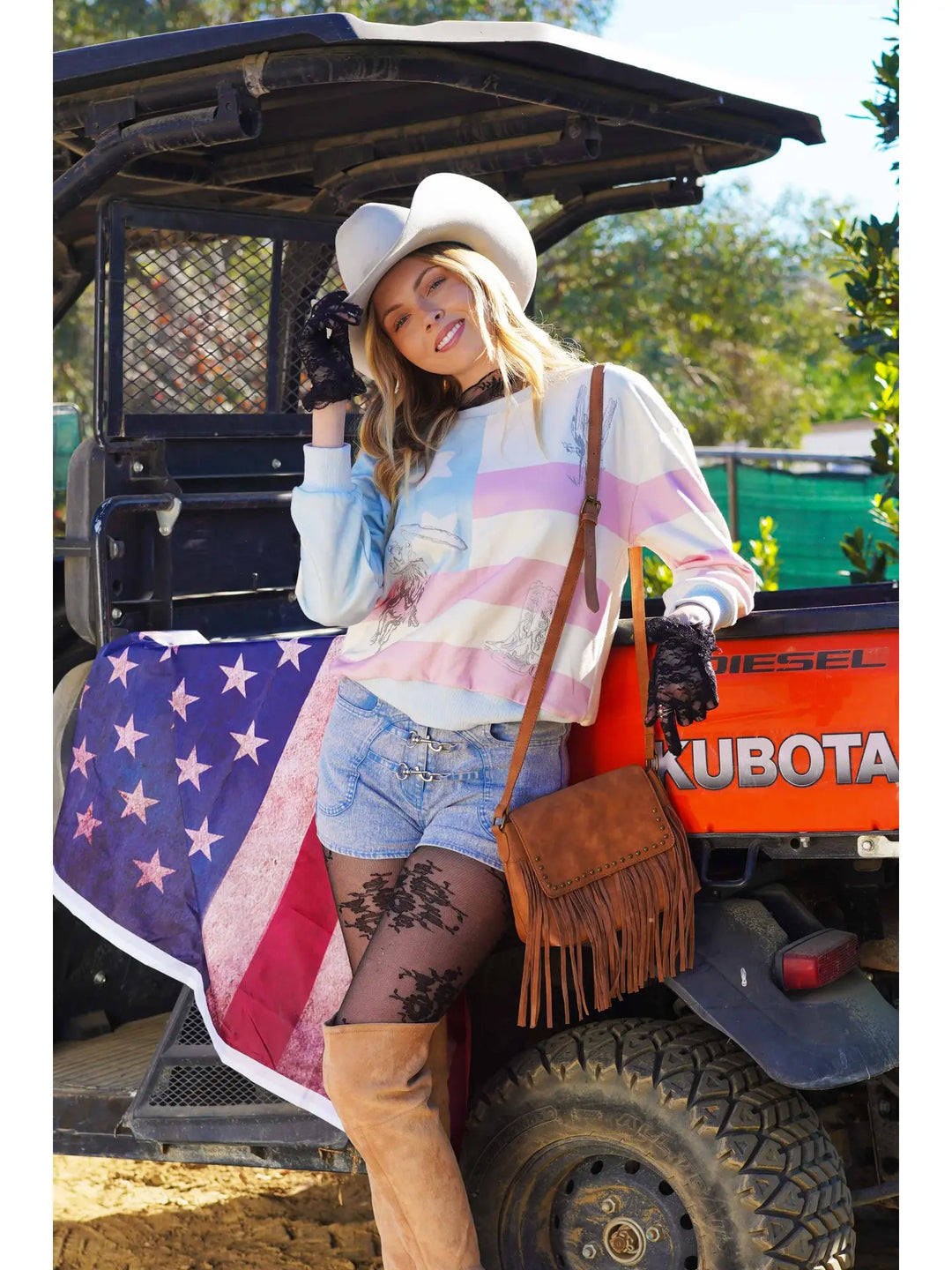 Woman in cowboy hat and sunglasses standing next to a vehicle with a large American flag draped over it.