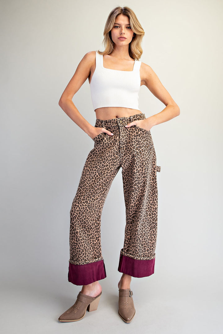 A model wearing leopard-print wide-leg pants with a contrasting deep purple cuff, styled with a white crop top and taupe heeled mule shoes against a light gray studio background.