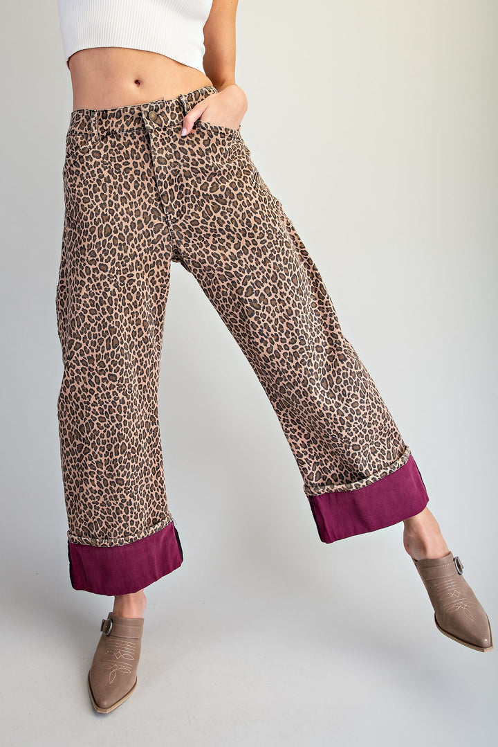 A model wearing wide-leg cropped pants with a brown and beige leopard print and a contrasting deep magenta hem, styled with brown ankle boots against a light gray studio background.