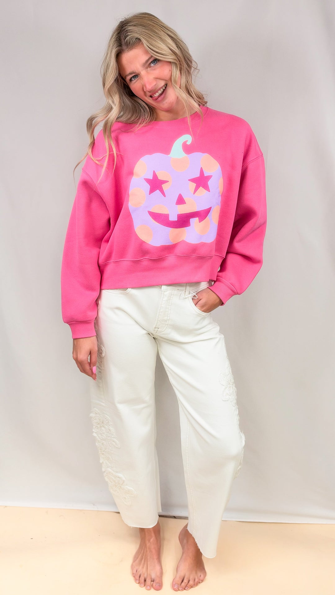 Woman wearing cream jeans with hot pink sweatshirt with purple polka dot pumpkin on front on plain background