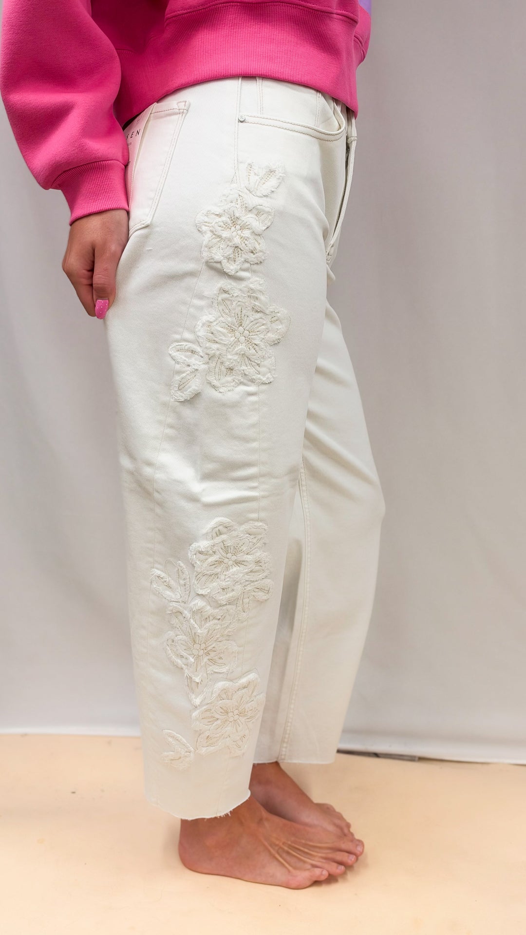 Woman wearing cream color jeans with flower appliques on side on plain background