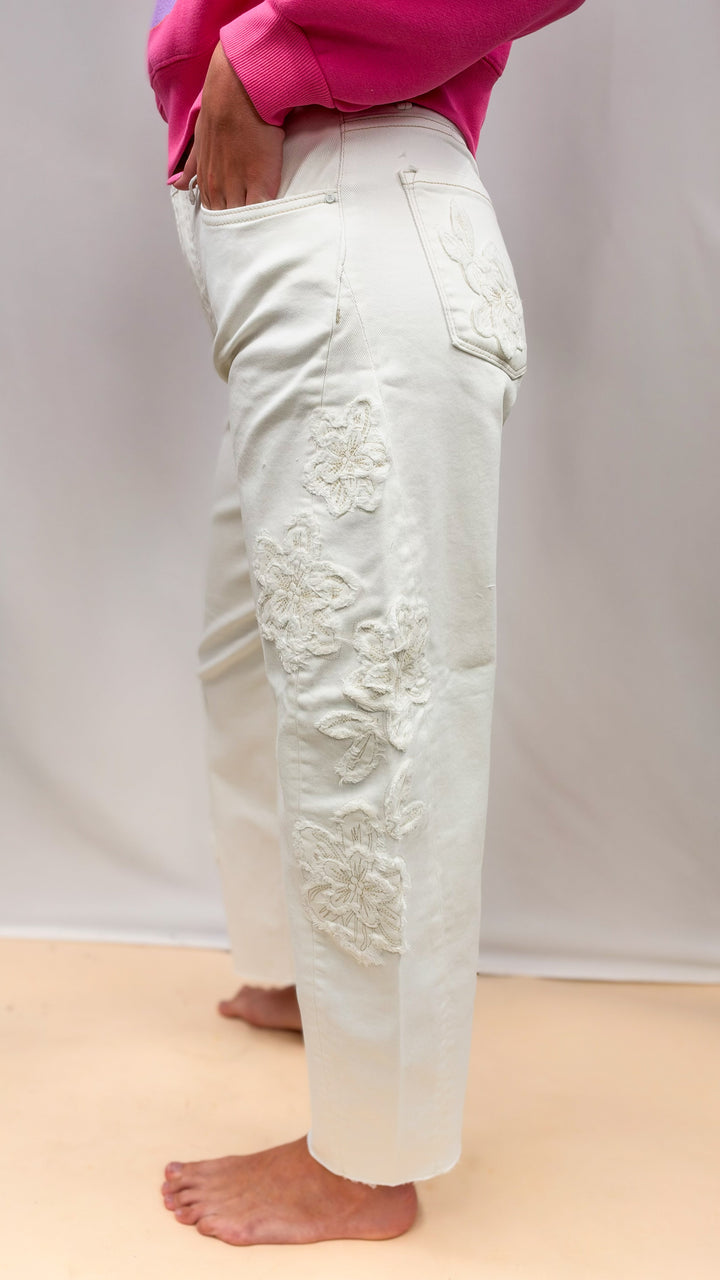 Woman wearing cream color jeans with flower appliques on side on plain background