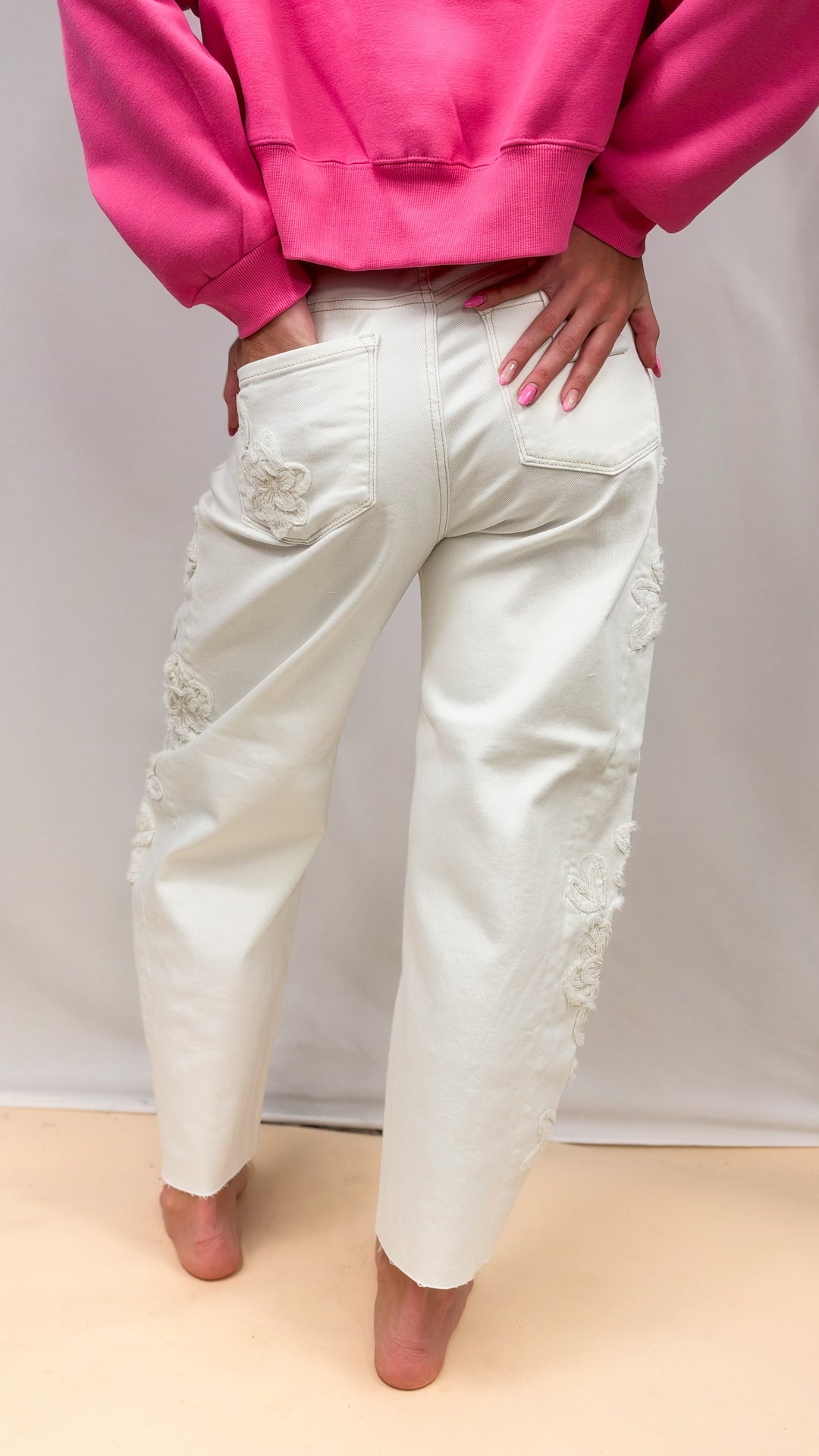 Woman shown from back wearing cream color jeans with flower appliques on side and pink sweatshirt on plain background