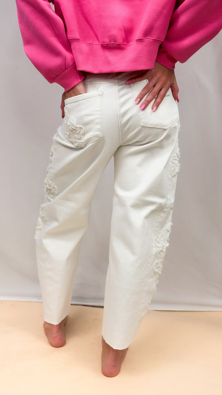 Woman shown from back wearing cream color jeans with flower appliques on side and pink sweatshirt on plain background