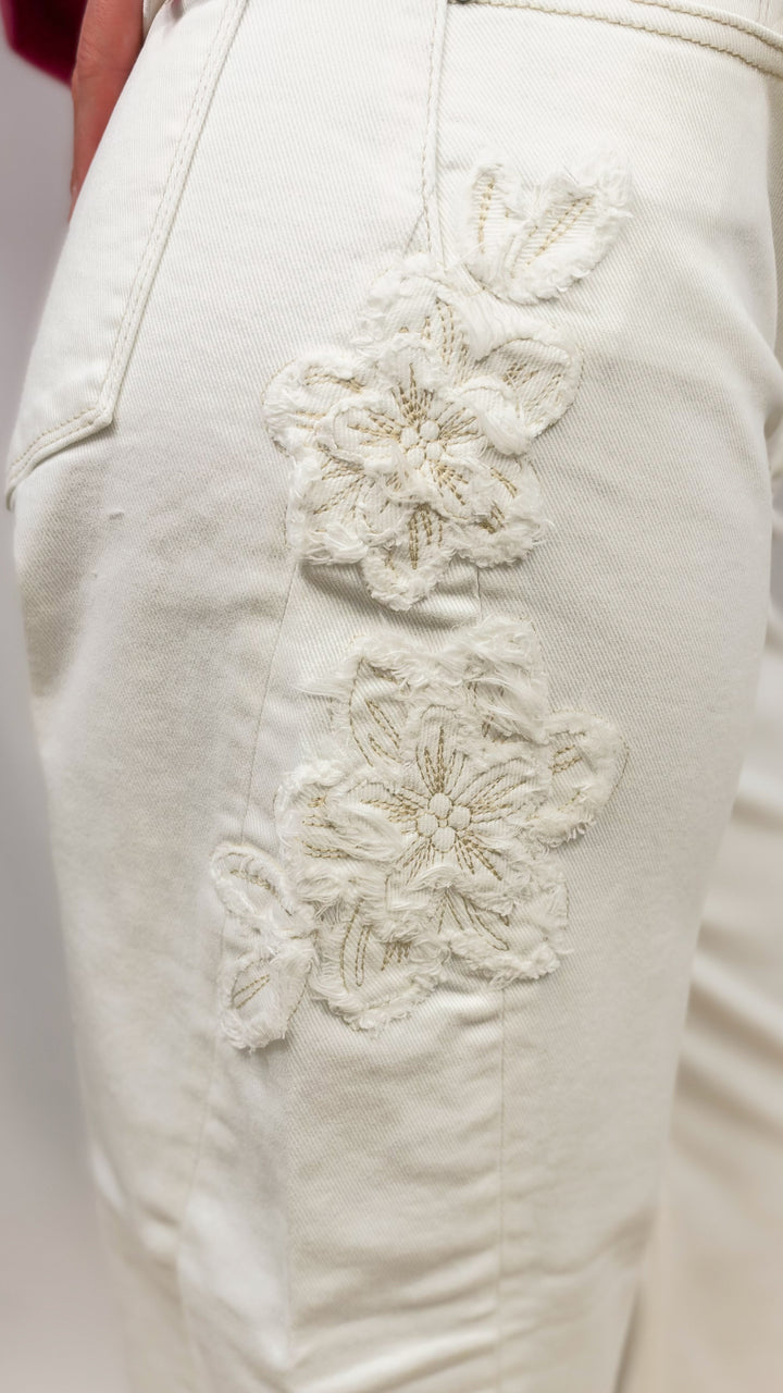 Woman wearing cream color pants showing up close detail shot of floral appliques on plain background