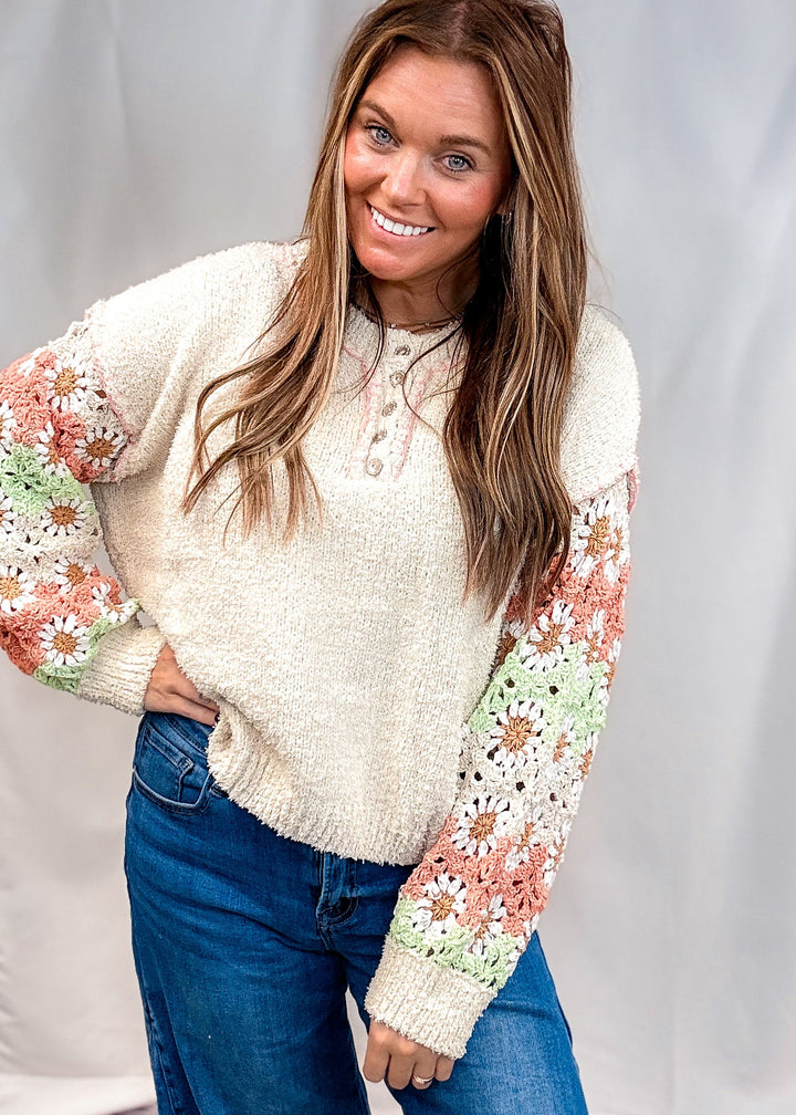 A woman modeling a cream-colored, fuzzy knit sweater with colorful floral crochet sleeve panels in pink, green, and white, paired with blue jeans against a white studio backdrop.