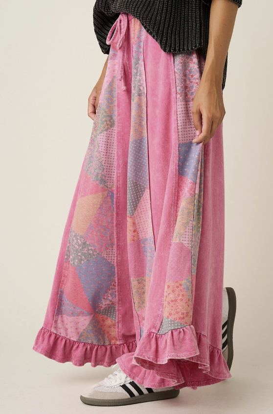 A person wearing a long pink patchwork skirt with ruffled hems, paired with white sneakers and a dark ribbed top, standing against a plain light beige studio background.