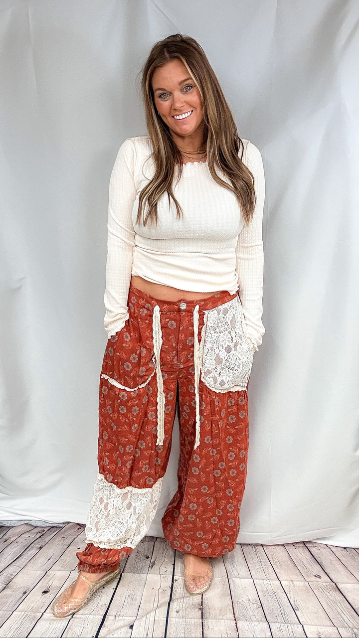 Woman wearing floral rust color pants with lace accents with cream shirt on plain background