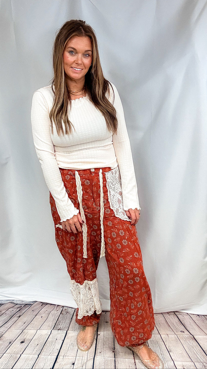 Woman wearing rust colored floral pants with cream ribbed top on plain background