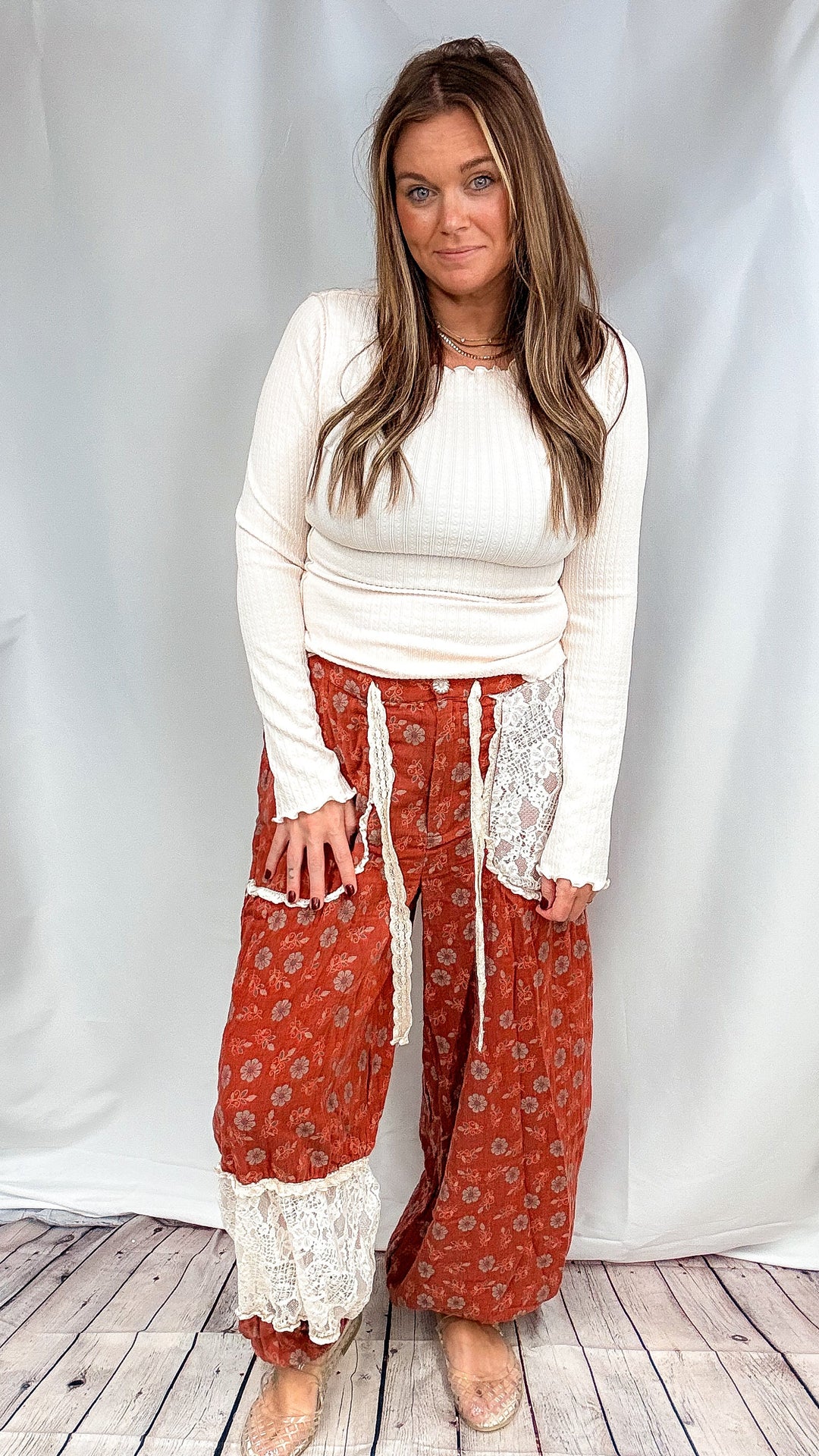 A woman modeling a white ribbed long-sleeve top and rust-orange patterned wide-leg pants with white lace trim, standing in front of a white fabric backdrop.