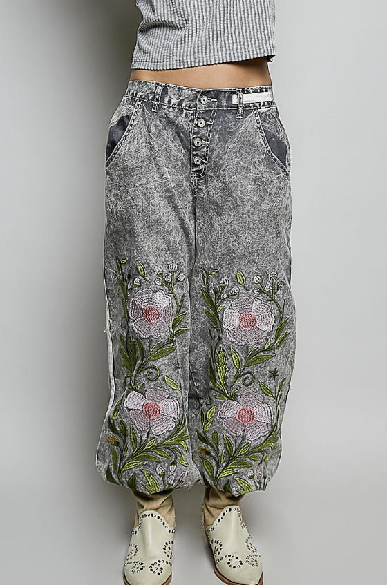 A person wearing acid-washed gray denim pants with large embroidered pink and white flowers and green leaves on the front, paired with cream western-style boots against a light gray studio background.
