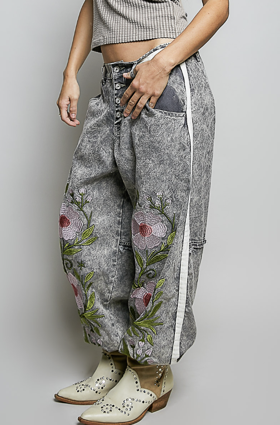 A person wearing gray acid-wash wide-leg jeans with pink and green floral embroidery on the lower legs, a white side stripe, and cream western-style boots, photographed against a light gray studio background.