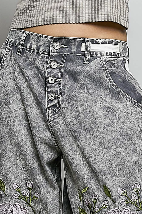 Close-up of gray acid-wash denim pants with a button fly, floral embroidery near the hem, and a striped cropped top partially visible at the top.
