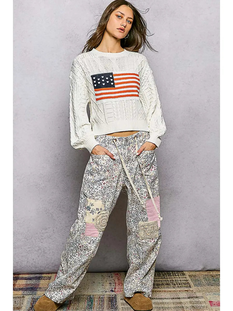 Woman wearing a white sweatshirt with an American flag design and patterned pants against a gray background.