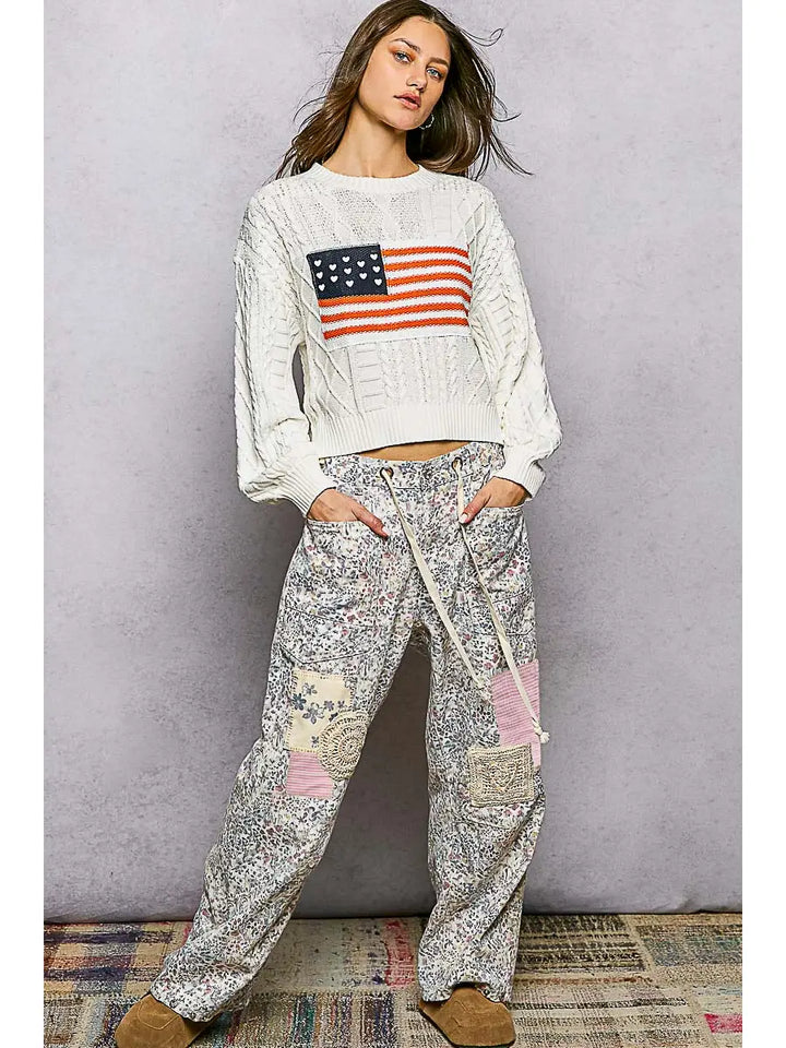 Woman wearing a white sweatshirt with an American flag design and patterned pants against a gray background.