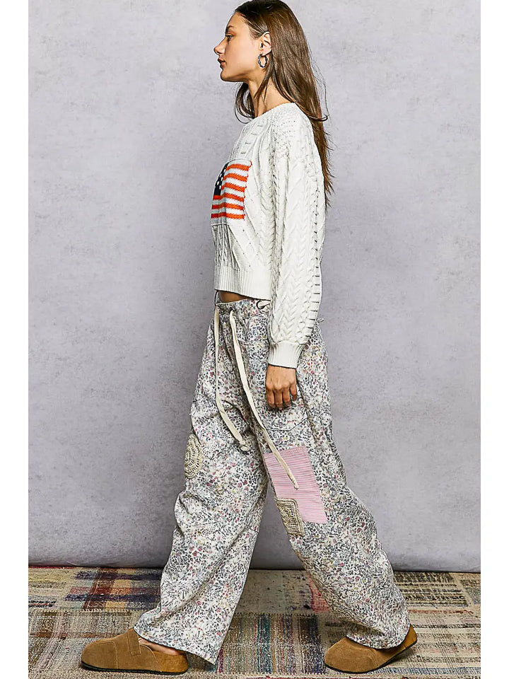 Woman wearing a white sweater with a colorful design and patterned pants against a plain background