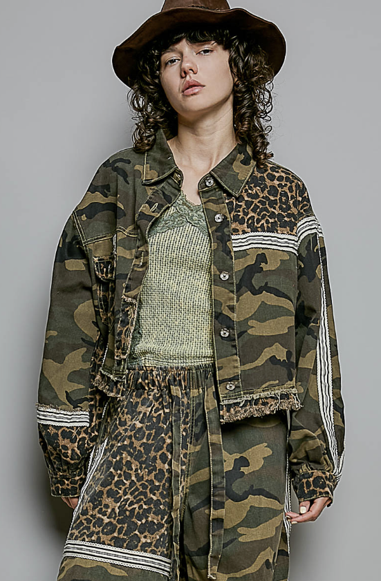 A model wearing a mixed-media camouflage and leopard print jacket with white stripe details, matching pants, a green knit top, and a brown wide-brim hat, photographed against a solid gray studio background.