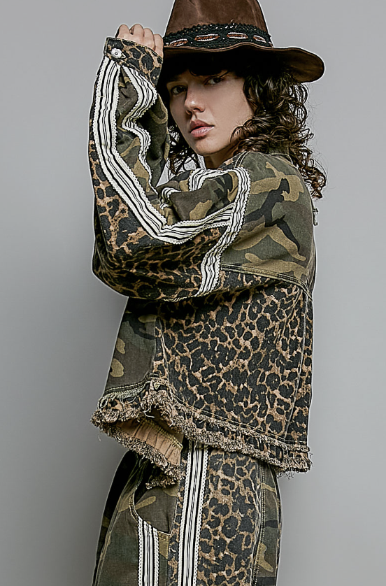 A model wearing a brown wide-brim hat, a distressed camouflage and leopard-print jacket with white stripe details, and matching pants, posed against a solid gray studio background.