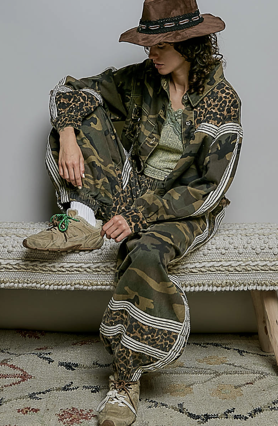 A person wearing a matching camouflage and leopard-print jacket and pants set with striped accents, a brown hat, and beige sneakers, seated on a woven bench against a plain wall.