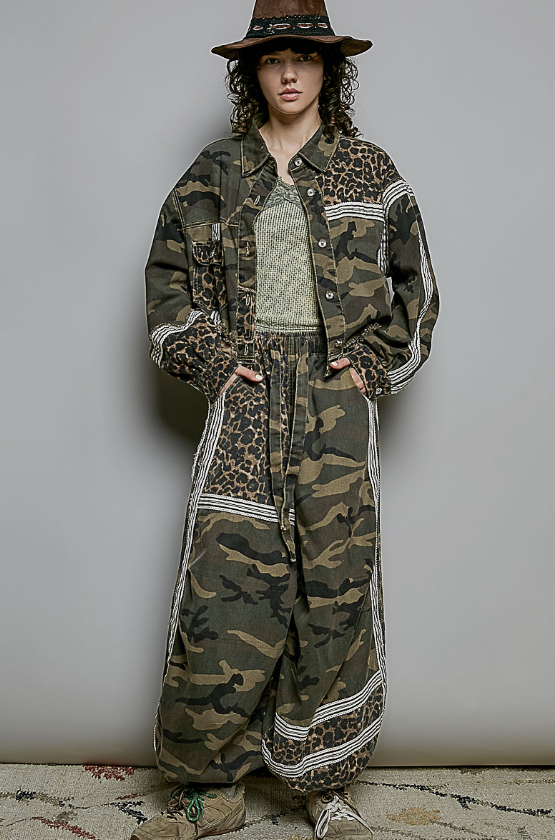 A model wearing a matching oversized two-piece outfit with a mixed print of leopard print and camouflage, styled with a brown wide-brim hat, a textured knit top, and sneakers against a plain gray wall.