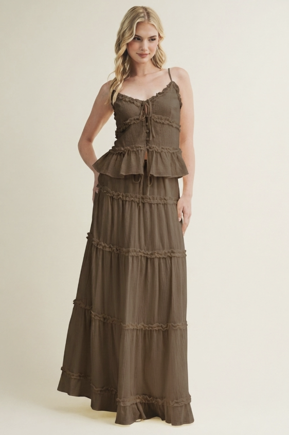 A woman modeling a sleeveless, tiered maxi dress in a warm taupe-brown color, standing against a solid light beige studio background.
