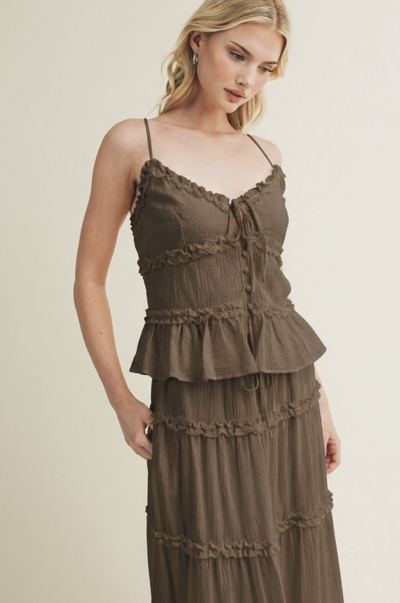 A woman wearing a brown ruffled spaghetti-strap dress with a tie-front bodice and tiered skirt, photographed against a plain light beige studio background.