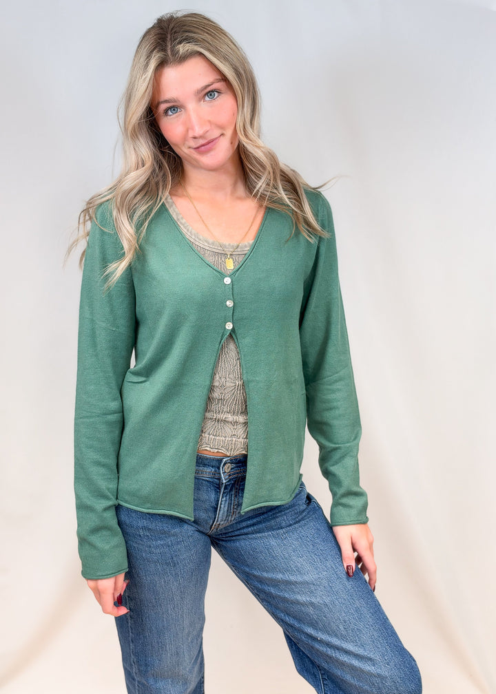 Woman wearing sage green 3 button cardigan with green tank and jeans on plain background