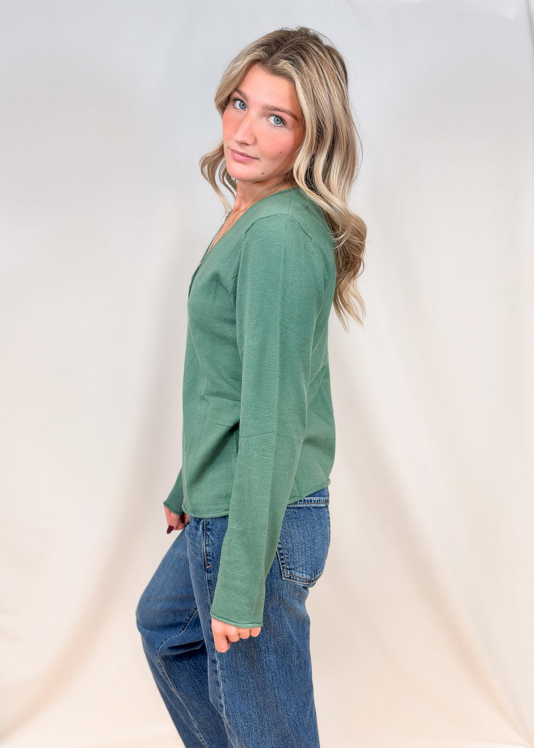 Woman shown from side view wearing sage green cardigan with jeans on plain background