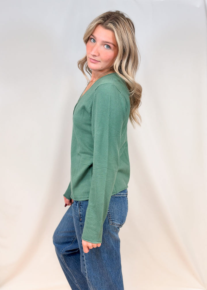 Woman shown from side view wearing sage green cardigan with jeans on plain background