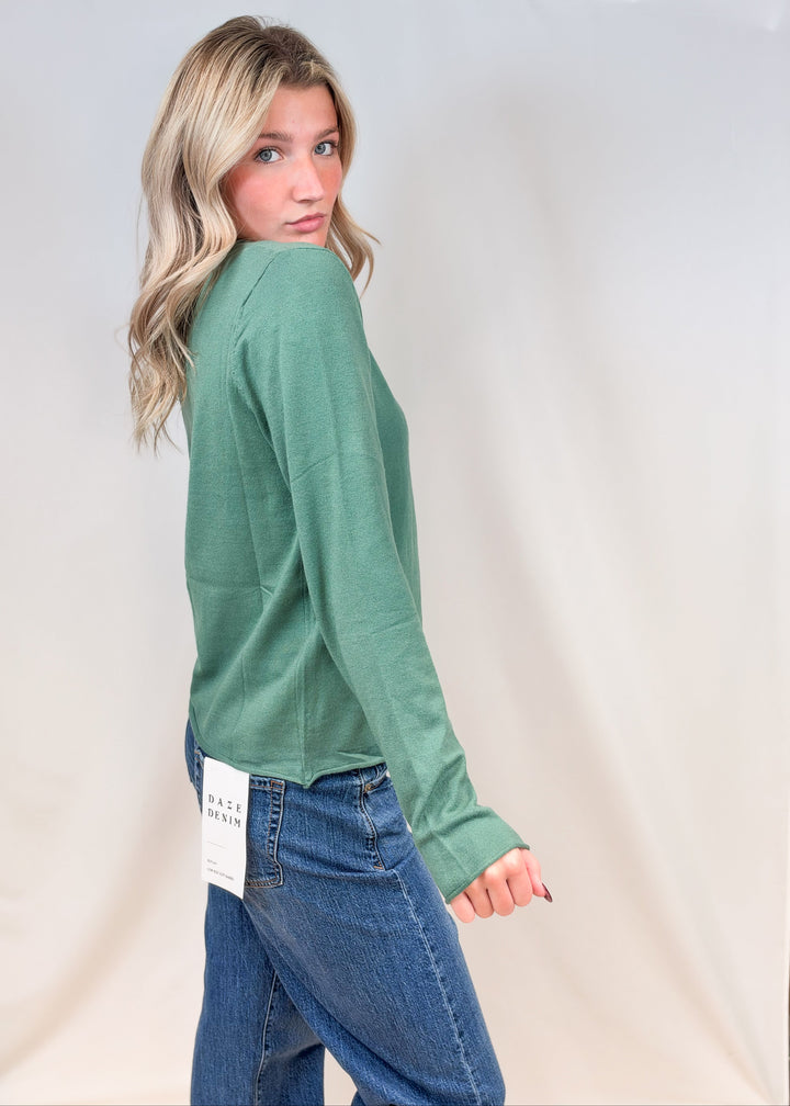 Woman shown from side view wearing sage green cardigan with jeans on plain background