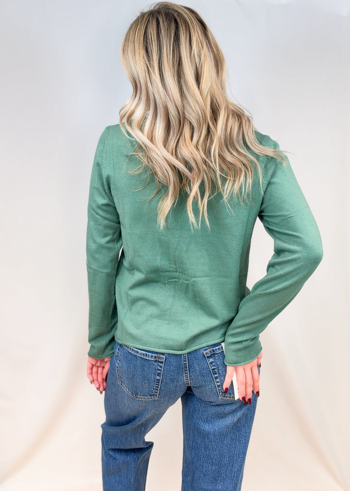 Woman shown from back view wearing sage green cardigan with jeans on plain background
