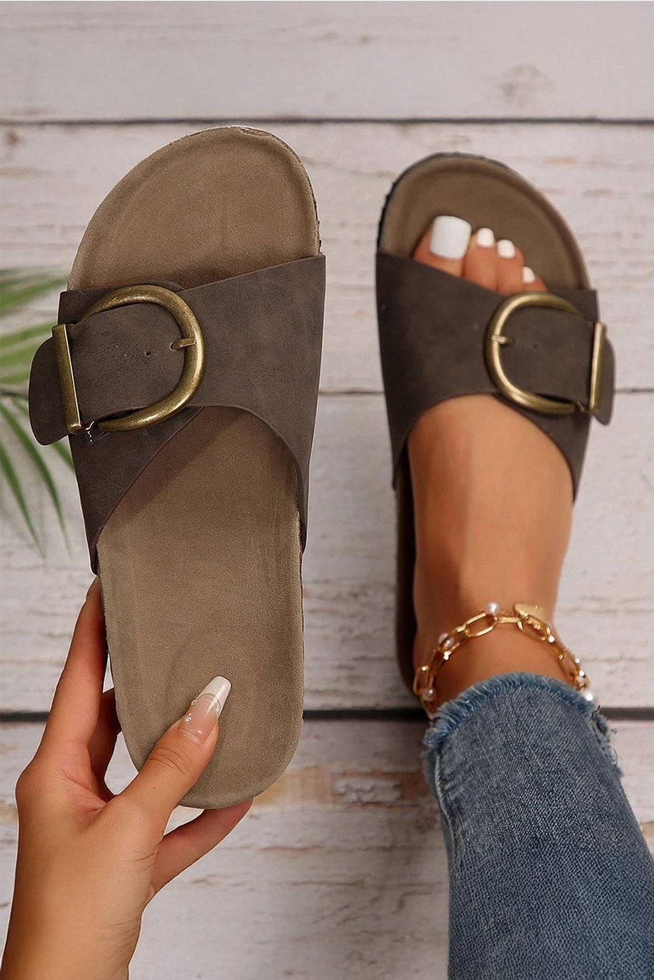 Brown sandals with gold buckles worn by a person on a wooden floor.