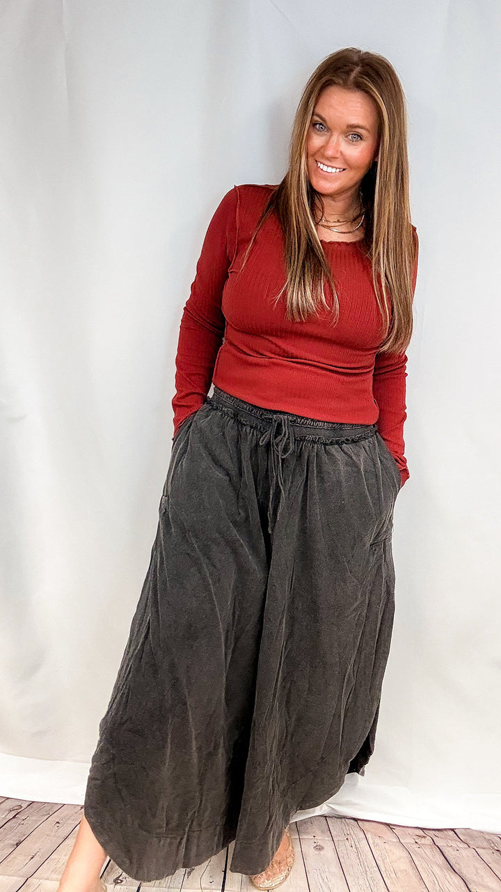 A woman modeling a red long-sleeve top and dark charcoal wide-leg linen pants against a white fabric backdrop.