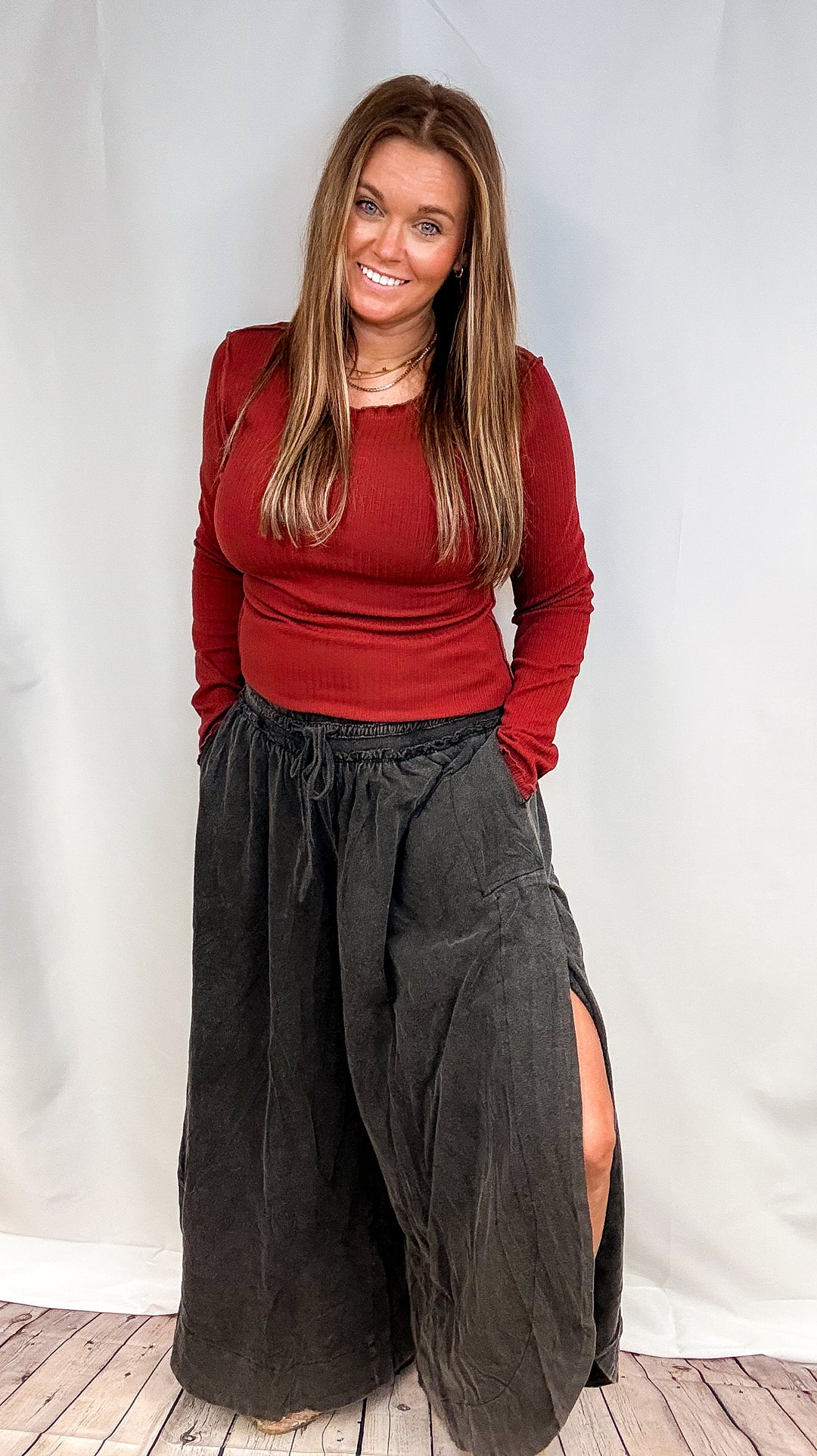 A woman modeling a dark red long-sleeve top and loose black wide-leg pants with a side slit, standing against a white studio backdrop.