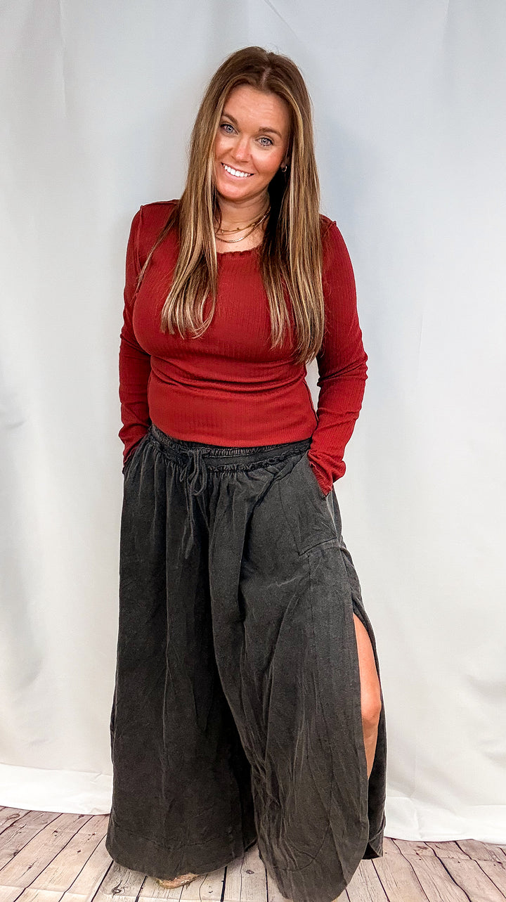 A woman modeling a dark red long-sleeve top and loose black wide-leg pants with a side slit, standing against a white studio backdrop.
