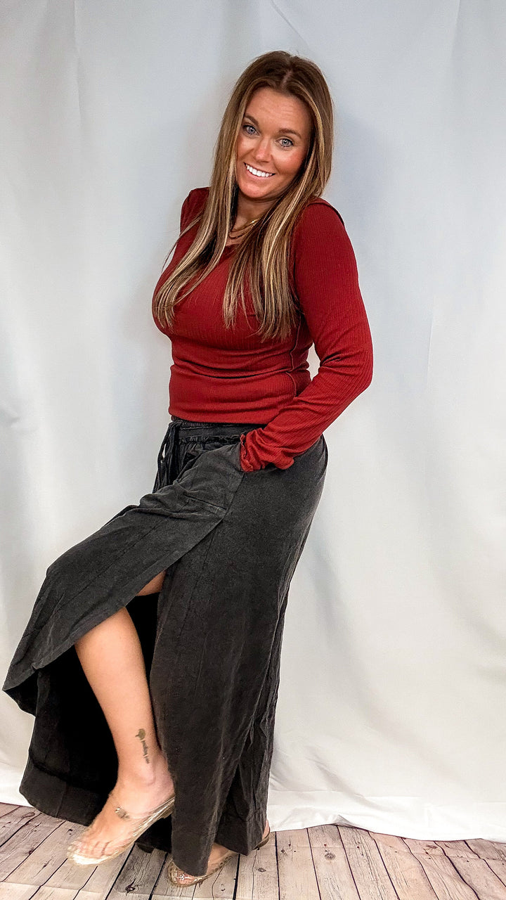 A woman posing in a studio setting wearing a long-sleeve red top and a dark gray high-low skirt with a side slit, standing against a white draped backdrop.