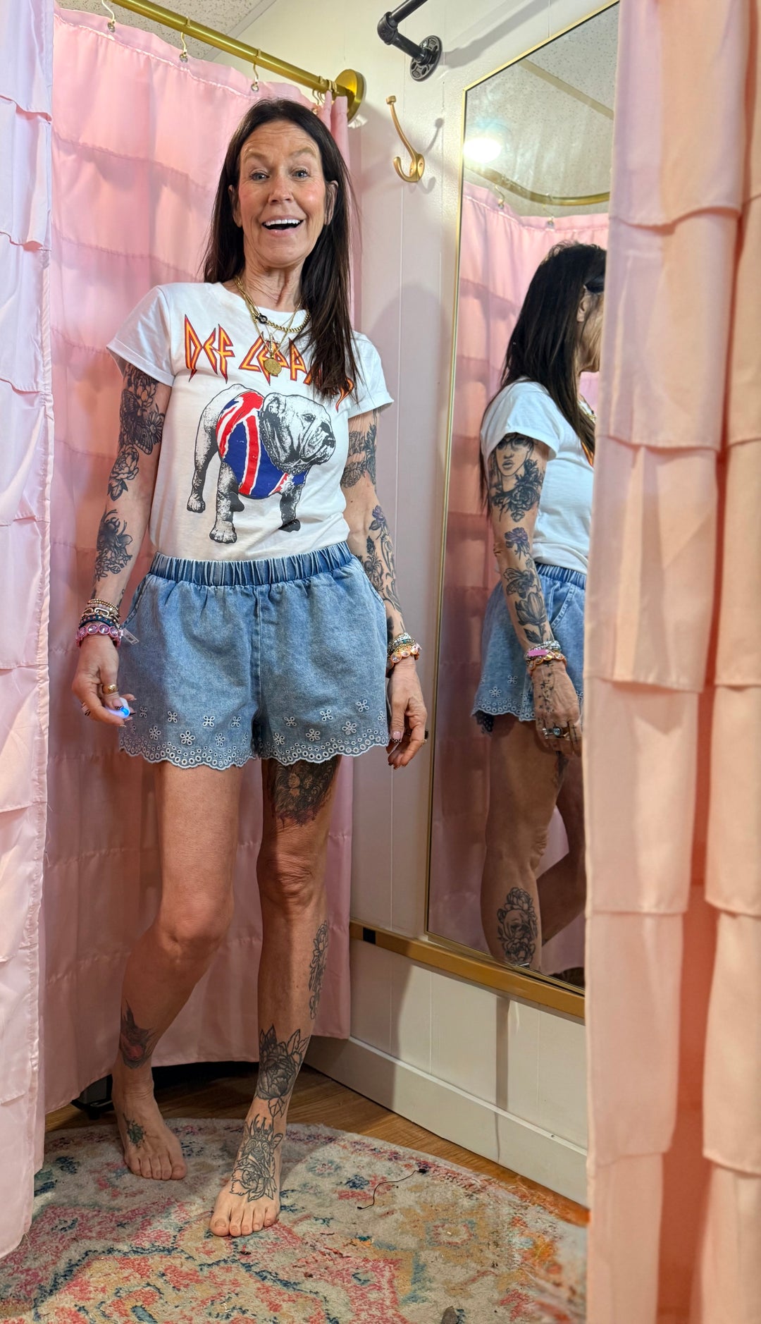 Person wearing a graphic t-shirt and denim shorts standing in front of a mirror with pink curtains.