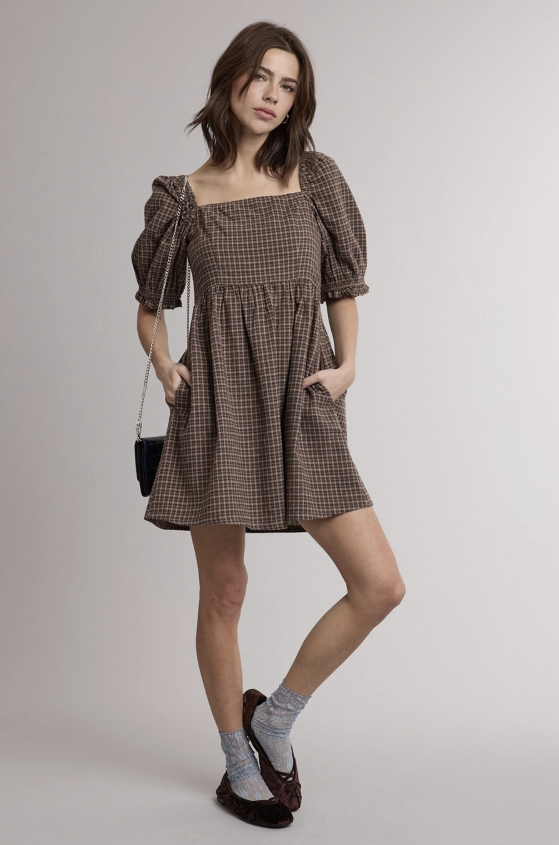 A woman models a brown and beige checkered puff-sleeve mini dress with a square neckline and side pockets, paired with gray socks and dark brown shoes against a light gray studio background.
