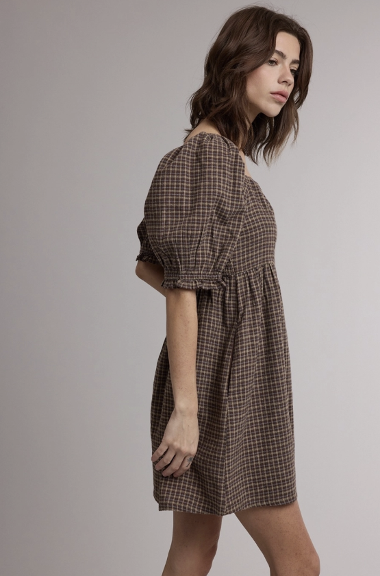 A woman modeling a brown and beige plaid puff-sleeve mini dress against a plain light gray studio background.