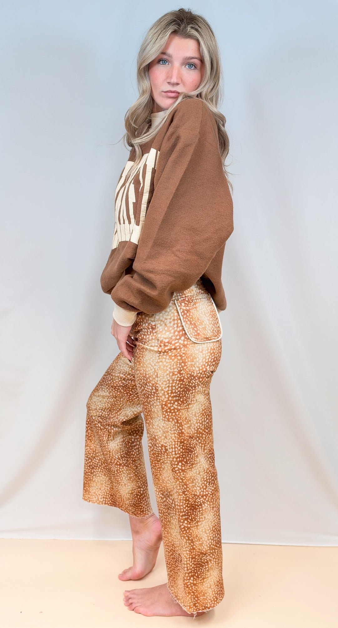 Woman shown from side view wearing brown mock neck sweatshirt with fawn print pants on plain background