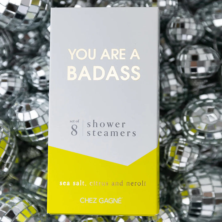 A rectangular product box for shower steamers stands upright against a background of mirrored disco balls. The box is white on the upper half with pale yellow text reading &