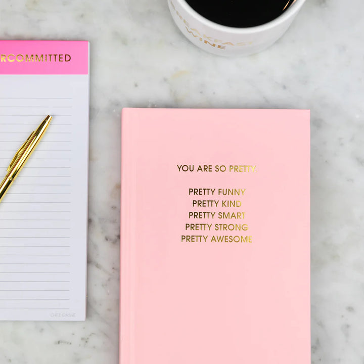 Pink notebook with gold text, white notepad, and coffee cup on marble surface