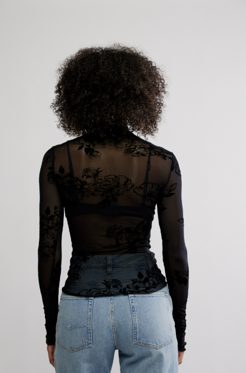 Woman shown from back wearing black mesh floral top with blue jeans on white background