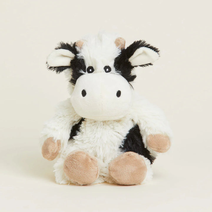 A plush cow toy with a fluffy white body, black patches, and beige hooves and ears, sitting against a plain light beige background.