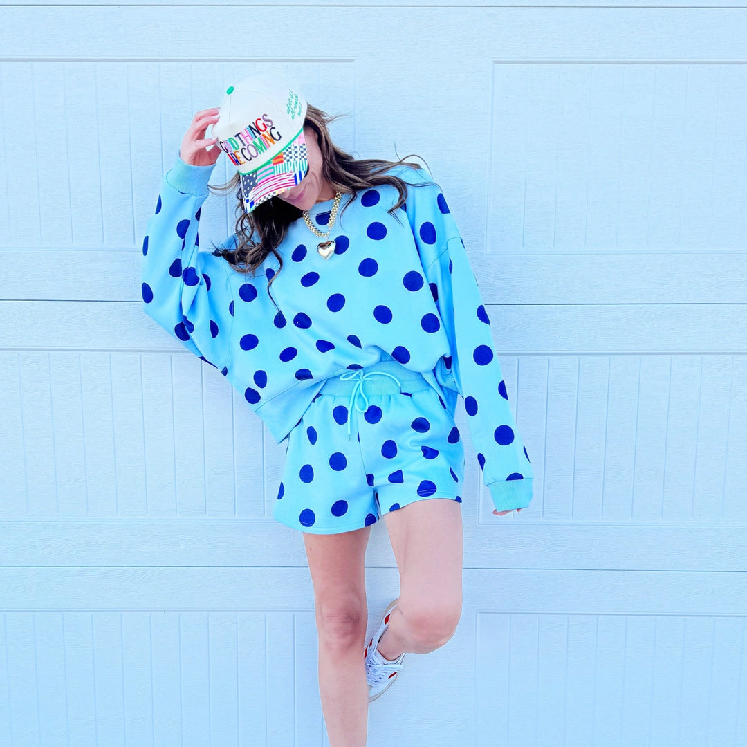 Person wearing a blue polka dot outfit with a colorful hat against a light blue tiled wall.
