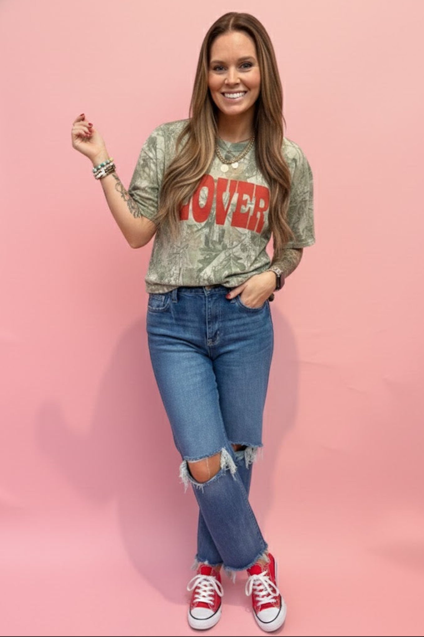 woman standing in front of pink wall wearing camo tshirt and jeans.
